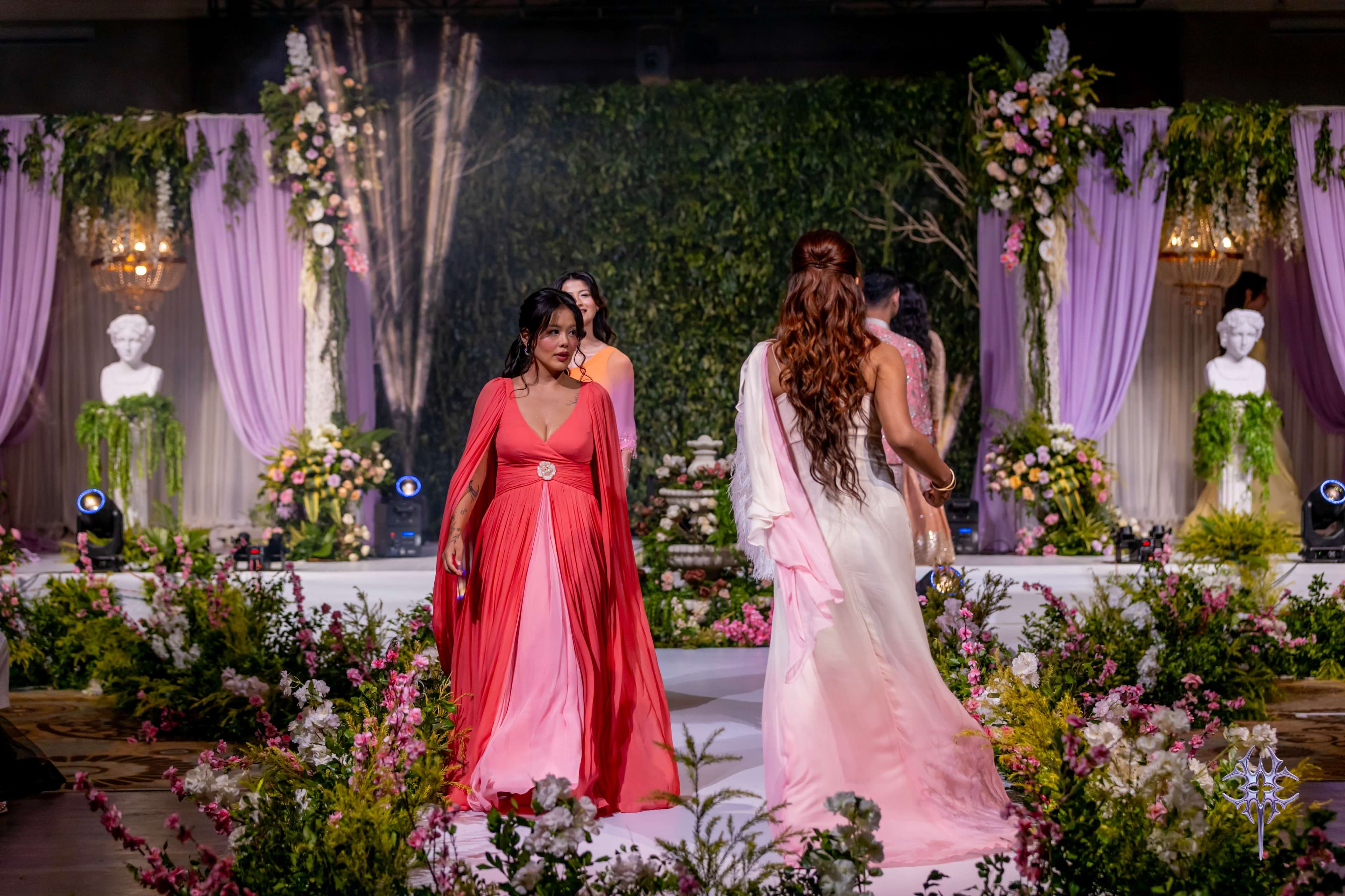 Two models on the Asteria runway in flowing coral and blush evening gowns, walking a flower-lined aisle